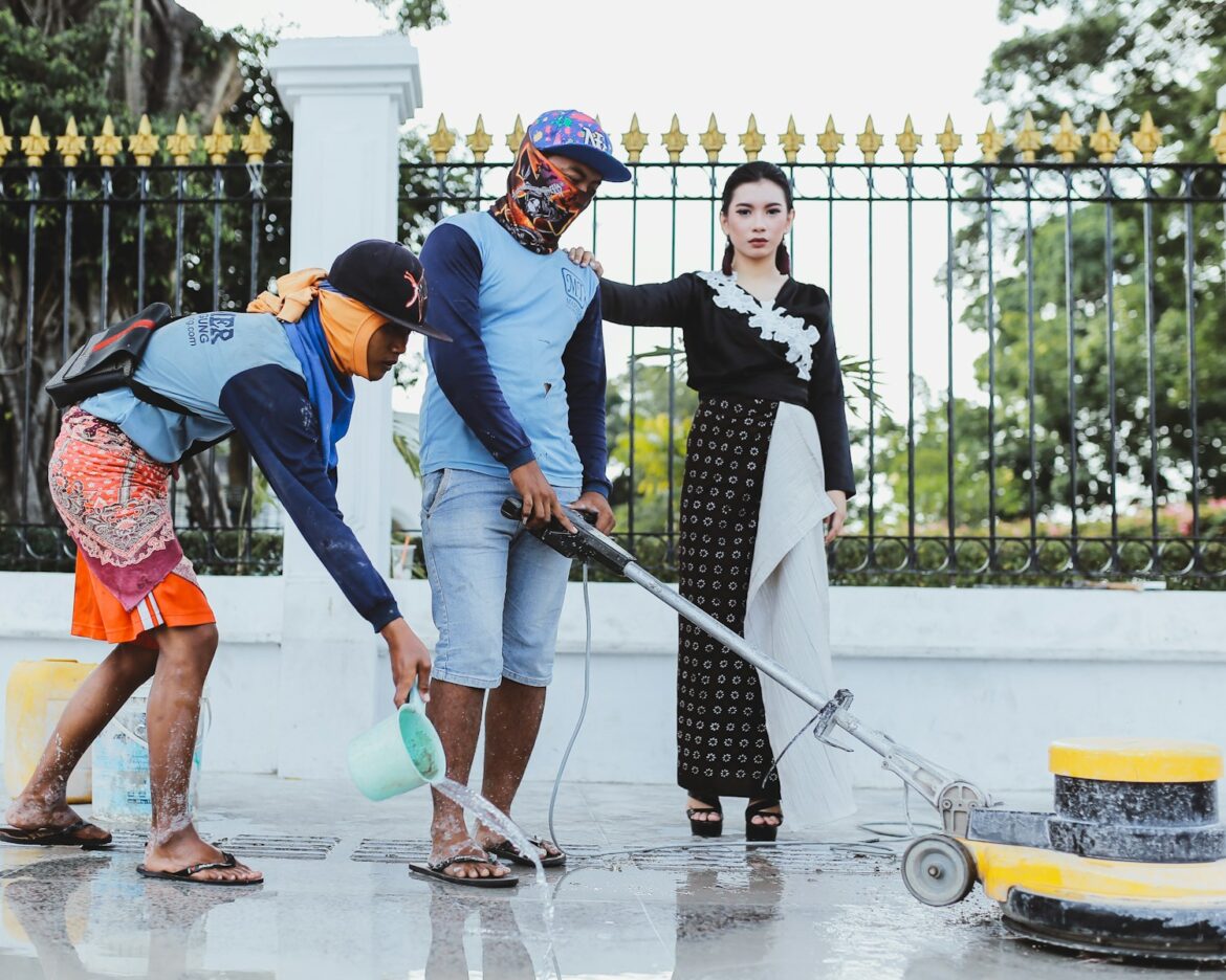 woman standing while holding on shoulder of man cleaning pathway and another man spreading water using green water dipper